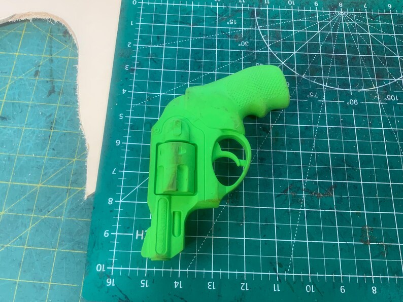 May include: A bright green toy revolver, resting on a green cutting mat with grid lines and degree markings. The toy gun is a detailed replica, featuring a handle, trigger, and cylinder. The background includes a piece of beige material.