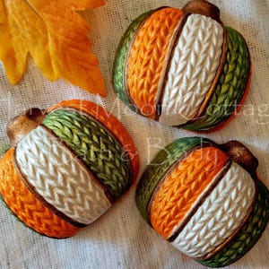 May include: Three decorative pumpkin-shaped items featuring a textured knit pattern. Each pumpkin displays alternating vertical segments in orange, white, and green with a metallic bronze-colored stem at the top. The surface has a raised cable knit design.