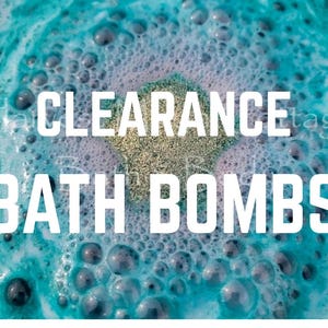 May include: A bath bomb dissolving in water, creating a vibrant turquoise and light purple foam with numerous bubbles. The text CLEARANCE BATH BOMBS is overlaid in large white capital letters across the center of the effervescent water surface.