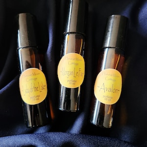 May include: Three perfume bottles with black caps and amber-colored glass are displayed. Each bottle has a yellow label with the text "Harvest Moon Cottage" and a perfume name: "Lady of the Lake", "Morgan Le Fay", and "Avalon". The labels also state "perfume 10 ml".