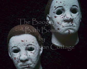 May include: Two white bath bombs shaped like the face of a horror movie character. The bath bombs are covered in red splatter and have black eyes. The text "The Boogeyman Bath Bomb by Harvest Moon Cottage" is written in white on a black background.