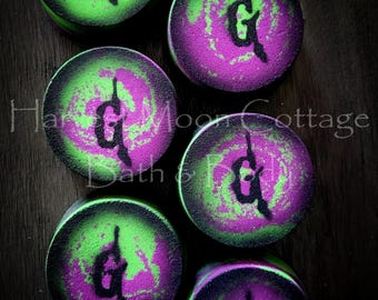 May include: Six circular bath bombs arranged in a grid, each featuring a vibrant swirl of neon green and deep purple colors. A stylized black letter G is printed in the center of each bath bomb. The text Harvest Moon Cottage Bath & Body is visible.
