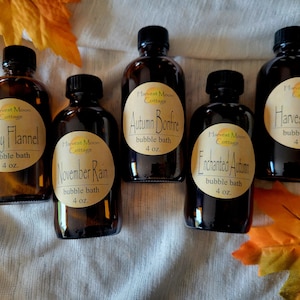 May include: Five amber glass bottles with labels for different bubble bath scents. The labels are brown with white text and feature a harvest moon logo. The scents are Cozy Flannel, November Rain, Autumn Bonfire, Enchanted Autumn, and Harvest Moon. Each bottle contains 4 ounces of bubble bath.
