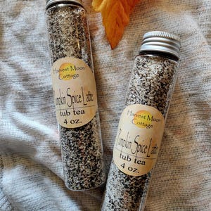 May include: Two clear glass tubes filled with a granular brown and black mixture, each topped with a silver screw cap. Each tube features a beige label reading Harvest Moon Cottage Pumpkin Spice Latte tub tea 4 oz. in black text.