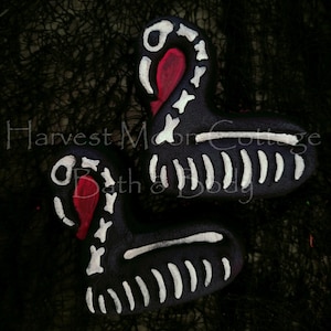 May include: Two black swan-shaped bath bombs with white skeleton detailing and red accents. The bath bombs are decorated with white paint to resemble bones. The text "Harvest Moon Cottage Bath & Body" is visible in the image.
