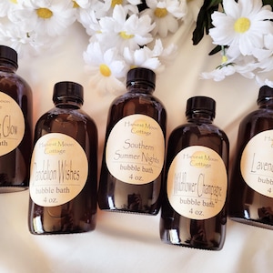 May include: Five amber glass bottles with labels that say "Harvest Moon Cottage" and different scents for bubble bath. The scents are "Lightning Bug Glow", "Dandelion Wishes", "Southern Summer Nights", "Wildflower Champagne", and "Lavender Honey". Each bottle contains 4 ounces of bubble bath.