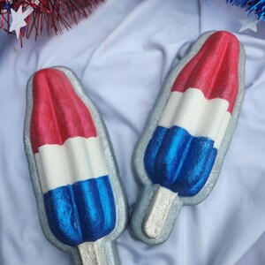 May include: Two red, white, and blue popsicle-shaped decorations with silver outlines and wooden sticks. The popsicles are arranged on a white fabric background, with red and blue tinsel in the background.