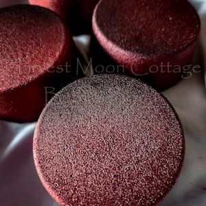 May include: Close-up of four round, dark red objects with a textured surface. The objects are arranged on a white fabric. The text "Harvest Moon Cottage Bath & Body" is partially visible.