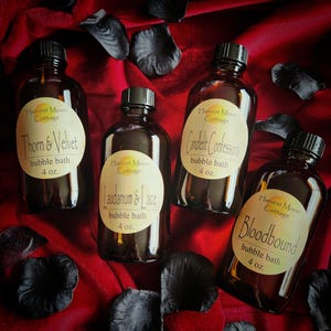 May include: Four dark glass bottles of bubble bath with black caps and cream-colored labels. The labels read "Harvest Moon Cottage" and feature names like "Thorn & Velvet" and "Bloodbound." Each bottle contains 4 oz of bubble bath.