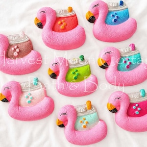 May include: A collection of pink flamingo-shaped bath bombs with colorful accents. Each flamingo has a yellow beak, black eye, and a decorative top with various colors like red, blue, green, and pink. The bath bombs are arranged on a white surface.