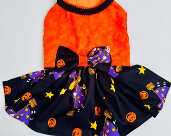 May include: A pet dress featuring a bright orange bodice with a black braided trim neckline. The skirt is black with a repeating pattern of orange jack-o'-lanterns, yellow stars, and purple-clad witches holding brooms. A matching bow is centered.