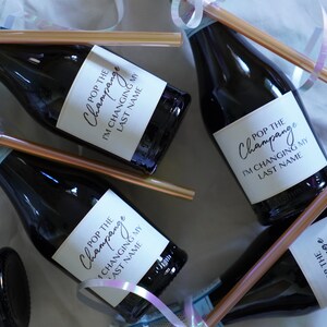 Könnte beinhalten: Eine Gruppe von Mini-Champagnerflaschen mit weißen Etiketten, auf denen "Pop the Champagne I'm Changing My Last Name" steht. Jede Flasche hat einen goldenen Strohhalm, der mit einem hellblauen Band um sie herum gebunden ist.