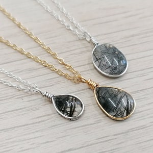 May include: Three necklaces with teardrop-shaped pendants. Each pendant features a clear stone with black inclusions, wire-wrapped in silver or gold. The chains are silver or gold, resting on a light wood-grain surface.