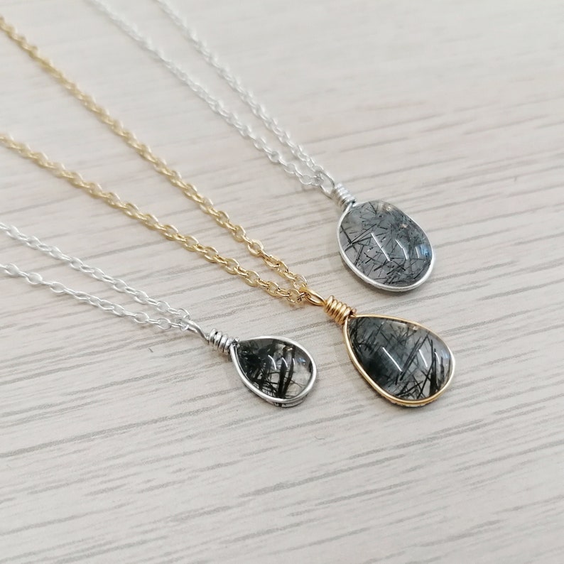 May include: Three necklaces with teardrop-shaped pendants. Each pendant features a clear stone with black inclusions, wire-wrapped in silver or gold. The chains are silver or gold, resting on a light wood-grain surface.