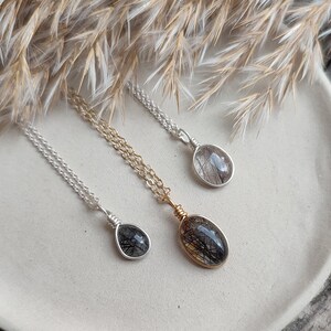 May include: Three necklaces with oval pendants. The pendants are clear with black inclusions, set in silver and gold wire. The chains are silver and gold. The necklaces are displayed on a white plate with dried grasses in the background.