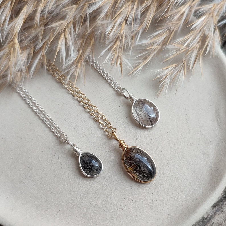 May include: Three necklaces with oval pendants. The pendants are clear with black inclusions, set in silver and gold wire. The chains are silver and gold. The necklaces are displayed on a white plate with dried grasses in the background.