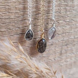 May include: Three necklaces with clear, oval-shaped pendants containing black inclusions. One pendant is wrapped in silver wire and hangs from a silver chain. The second pendant is wrapped in gold wire and hangs from a gold chain. The third pendant is wrapped in silver wire and hangs from a silver chain.