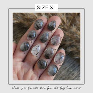 May include: A hand displays eleven XL-sized, polished gemstones. Each stone is a different shape and features black inclusions within a clear or translucent base. The text "SIZE XL" is at the top, and "choose your favourite stone from the drop down menu!" is at the bottom.