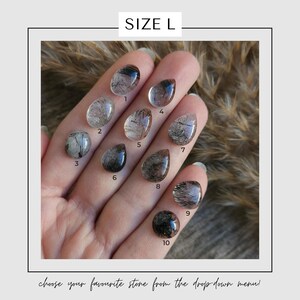 May include: A hand displays ten polished, clear quartz stones with black inclusions, each numbered. The stones vary in shape, including oval and teardrop. The text "SIZE L" is at the top, and the text "choose your favourite stone from the drop down menu!" is at the bottom.