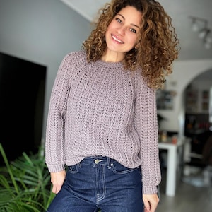 Op de afbeelding: Een lichtpaarse trui met lange mouwen en een textuurgebreid patroon. De trui wordt gecombineerd met een donkerblauwe spijkerbroek. Het model lacht, met krullend bruin haar.