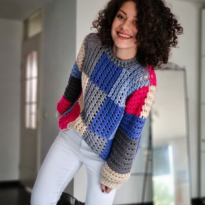 May include: A colorful crocheted sweater with a patchwork design in shades of blue, pink, beige, and gray. The sweater has a loose fit and a round neckline.