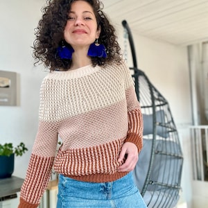 Peut inclure: Une femme portant un pull rayé marron, beige et blanc à manches longues. Elle porte une jupe en jean bleu et des boucles d'oreilles en forme de fleurs bleues.