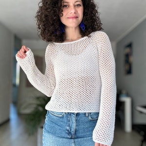 Könnte beinhalten: Ein weißer Häkelpullover mit lockerer Passform und langen Ärmeln. Der Pullover hat ein zartes Lochmuster.