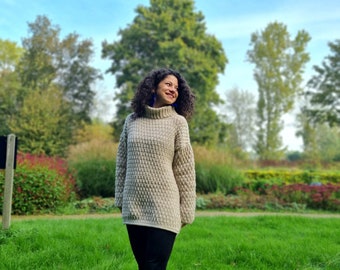 May include: A beige, chunky knit turtleneck sweater. The sweater has a textured pattern and long sleeves. The person is wearing black pants and black boots. The background includes green grass, trees, and a blue sky.
