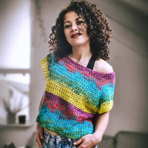 Puede incluir: Una mujer lleva una blusa de crochet colorida con un patrón de rayas de arcoíris. La blusa es de hombros descubiertos y tiene un corte holgado. La mujer lleva pantalones vaqueros azules.