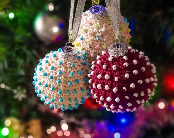 Op de afbeelding: Drie gehaakte kerstballen in wit, rood en crème met witte kralen. De ballen hangen aan een boom.