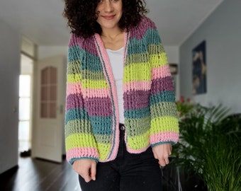 Puede incluir: Un cárdigan de crochet a rayas con rayas rosas, verdes y moradas. El cárdigan tiene un ribete blanco alrededor de los bordes y está abierto en la parte delantera.