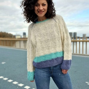 May include: A white crocheted sweater with a blue and green stripe at the bottom. The sweater has a loose fit and a round neckline.