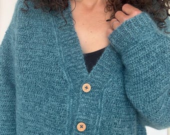 Op de afbeelding: Een teal-kleurig, oversized vest met een V-hals en twee houten knopen. De trui heeft een textuurgebreid ontwerp en lange mouwen. De ronde knopen hebben een kruisvormig ontwerp. Een gezellig, casual kledingstuk.