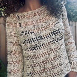 May include: A beige, open-weave crochet sweater with long sleeves. The sweater has a round neckline and a textured, see-through design. The crochet pattern creates a delicate, airy look. The garment is made of a light-colored yarn.