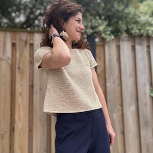 Op de afbeelding: Een beige gehaakte top met korte mouwen, een ronde halslijn en een rechte zoom, gecombineerd met een donkerblauwe broek. De top heeft een consistent, strak steekpatroon over het gehele oppervlak, wat zorgt voor een gestructureerde uitstraling.