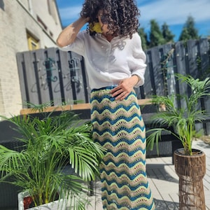 Puede incluir: Una mujer con una camisa blanca y una falda larga de ganchillo con un patrón ondulado en tonos verdes, azules y marrones. La falda es de largo midi y tiene un corte holgado.