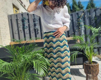 May include: A woman wearing a white shirt and a long, crocheted skirt with a wavy pattern in shades of green, blue, and brown. The skirt is a midi length and has a loose fit.