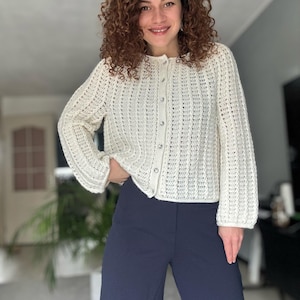 May include: A cream-colored, button-down cardigan with a textured knit pattern. The long-sleeved sweater is paired with navy blue wide-leg pants. The cardigan has silver-colored buttons.
