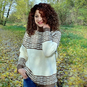 Könnte beinhalten: Ein weißer und brauner Häkelpullover mit geometrischem Muster. Der Pullover hat einen hohen Kragen und lange Ärmel. Die Person, die den Pullover trägt, steht in einer Waldlandschaft.