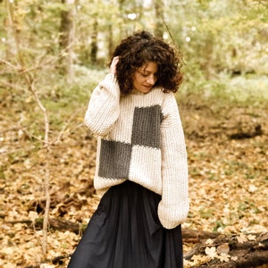 May include: A cream-colored sweater with a large gray and cream checkered pattern. The sweater has a relaxed fit and long sleeves.