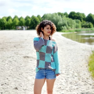 Könnte beinhalten: Eine Frau trägt ein blaues und graues kariertes Häkeltop mit langen Ärmeln und einer lockeren Passform. Sie steht auf einem Sandweg mit einem Gewässer im Hintergrund.