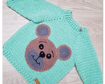 May include: A light green crocheted sweater with a brown bear face appliqué. The bear has gray ears, a gray nose, and black eyes.