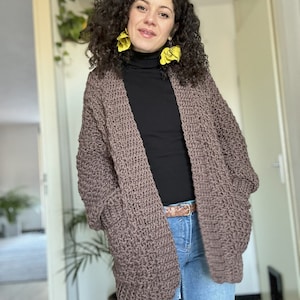 May include: A brown knitted cardigan with a black turtleneck shirt and blue jeans. The cardigan has pockets on the sides and a loose fit.