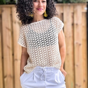 May include: A white crochet top with a loose, open weave pattern. The top has short sleeves and a round neckline. The model is wearing white shorts and has curly brown hair.