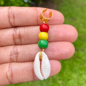 May include: Gold-toned ear cuff with a red, yellow, and green beaded accent, and a white seashell charm. The beads are arranged vertically, with the seashell hanging below. The cuff is held in a hand against a blurred green background.