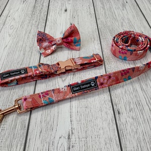 May include: A matching dog collar, leash, and bow tie set. The set features a floral pattern in shades of peach, pink, and blue. The collar and leash have rose gold hardware. The bow tie is pre-tied. The brand name "Abies Boutique" is printed on the collar and leash.