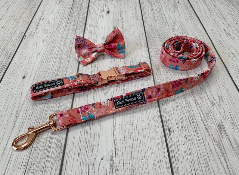 May include: A matching dog collar, leash, and bow tie set. The set features a floral pattern in shades of peach, pink, and blue. The collar and leash have rose gold hardware. The bow tie is pre-tied. The brand name "Abies Boutique" is printed on the collar and leash.