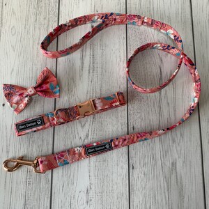 May include: A floral dog collar, leash, and bow tie set. The set features a pink and white floral pattern with blue accents. The collar has a rose gold buckle, and the leash has a matching rose gold clasp. The bow tie is pre-tied.