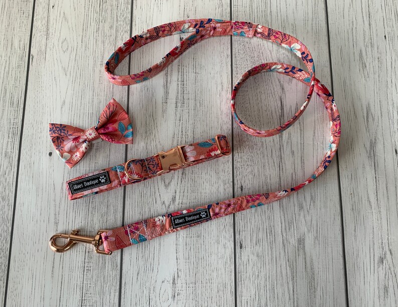 May include: A floral dog collar, leash, and bow tie set. The set features a pink and white floral pattern with blue accents. The collar has a rose gold buckle, and the leash has a matching rose gold clasp. The bow tie is pre-tied.