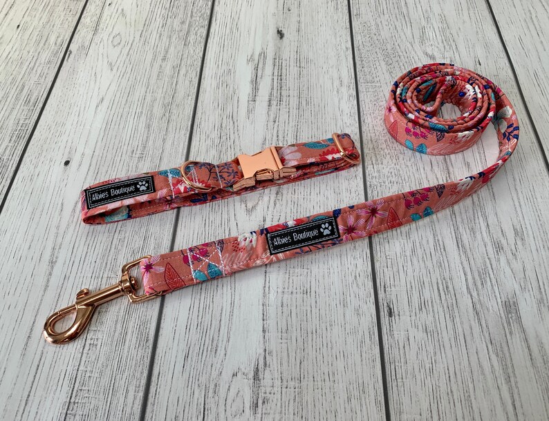 May include: A dog collar and leash set with a floral pattern in shades of peach, pink, and blue. The collar has a rose gold buckle, and the leash has a matching rose gold clasp. The set includes the text "Aloes Boutique".